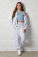 Woman wearing a NorthByOne Olive Zipper Hoodie in Marle Grey over a blue crop top with matching sweatpants standing against a white brick wall