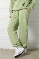 Person wearing the NorthByOne Emma Baggy Sweat Pant in Avocado, with elasticated waistband and side pockets standing against white brick wall