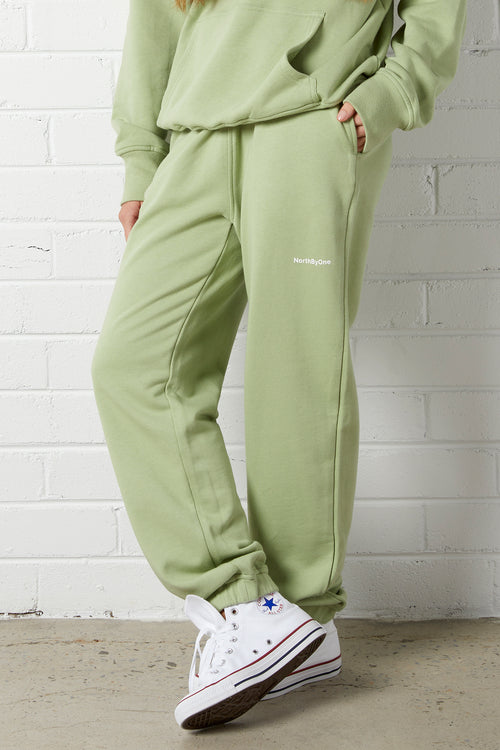 Person wearing the NorthByOne Emma Baggy Sweat Pant in Avocado, with elasticated waistband and side pockets standing against white brick wall