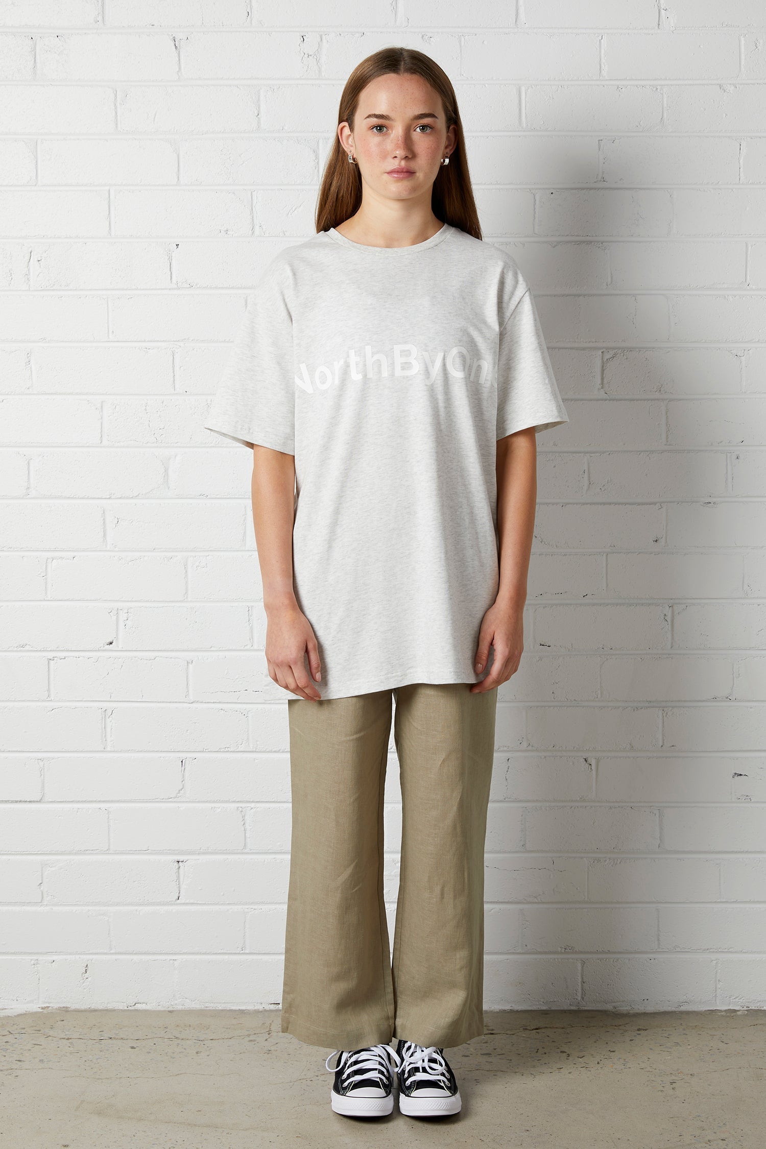 Woman wearing Lucy Oversized Tee | NorthByOne in Marle Grey with khaki pants and black sneakers standing against white brick wall