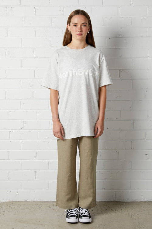 Woman wearing Lucy Oversized Tee | NorthByOne in Marle Grey with khaki pants and black sneakers standing against white brick wall