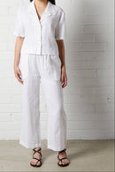 Woman wearing NorthByOne Sadie Linen Pant in white with a short-sleeve button-up shirt standing against a white brick wall