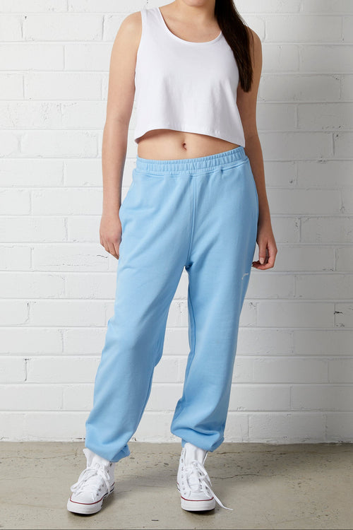 Woman wearing a NorthByOne Allegra Crop singlet in a white colour with a blue pair of sweats and white sneakers standing against a white brick wall
