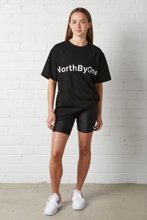 Young woman wearing Lucy Oversized Tee | NorthByOne in black with black bike shorts and white sneakers against white brick wall