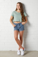 Young woman wearing a green cropped t-shirt and NorthByOne Charlotte Denim Short standing against a white brick wall