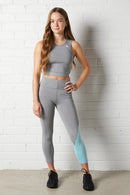 Young woman wearing the NorthByOne Cara Three Quarter Tight - Marle Grey Aqua and a grey sleeveless crop top standing against a white brick wall